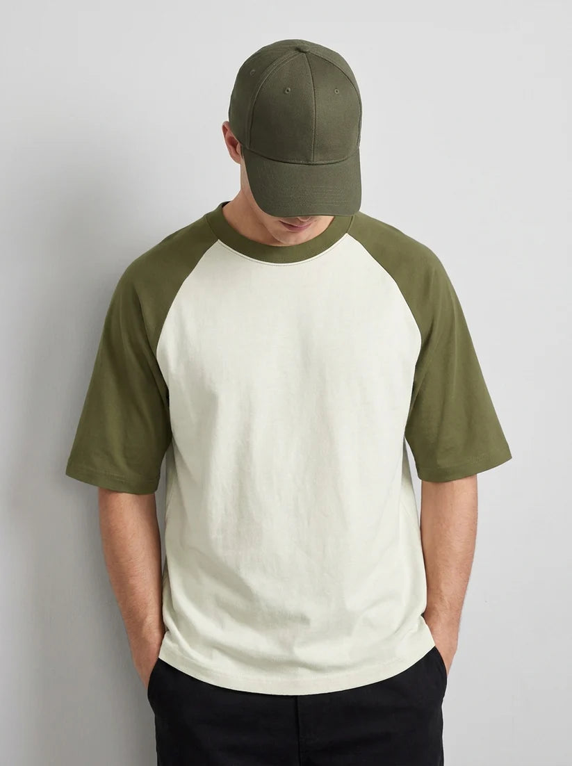 A man wearing a two-tone raglan t-shirt with an off-white body and olive green sleeves, paired with an olive green baseball cap and dark pants, standing against a light gray wall.