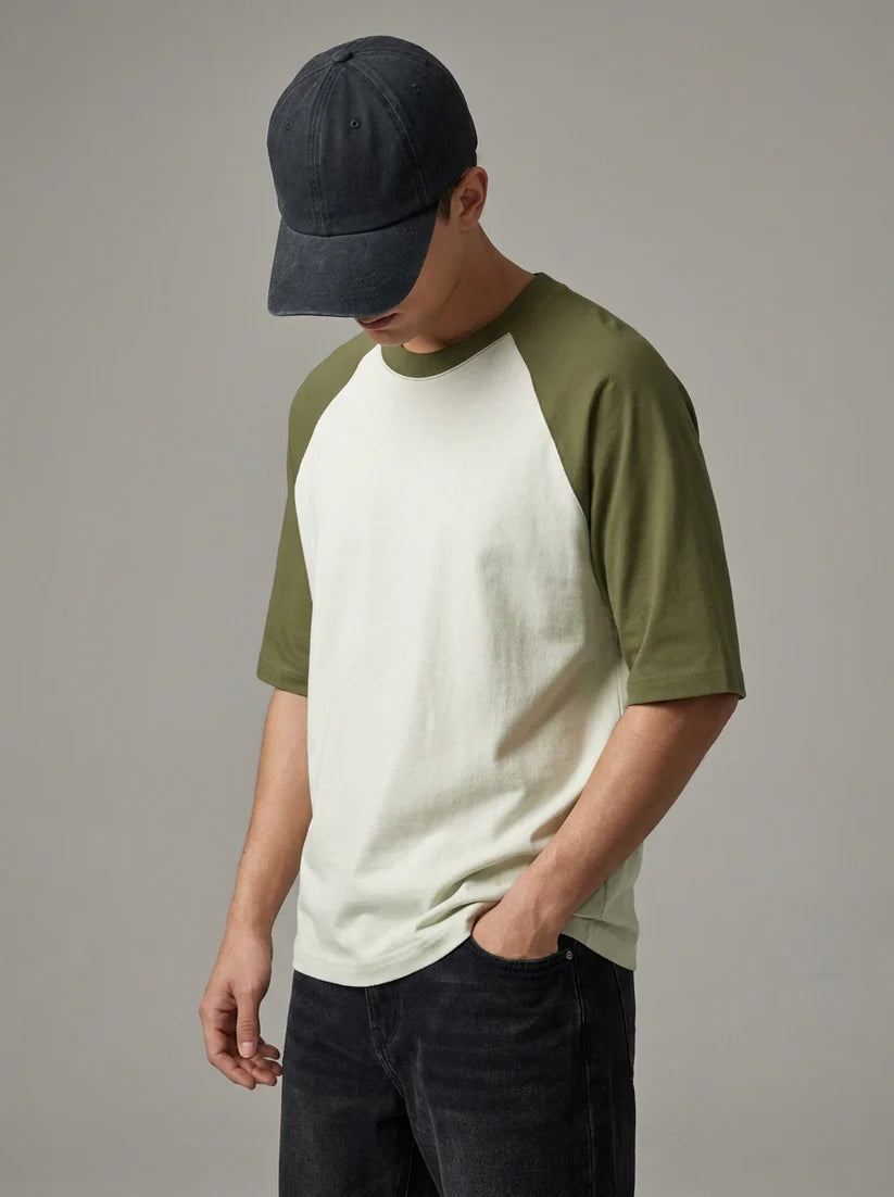A man wearing a raglan-style short-sleeve t-shirt with an off-white body and olive green sleeves, paired with a dark navy baseball cap and dark jeans, photographed against a neutral gray studio background.