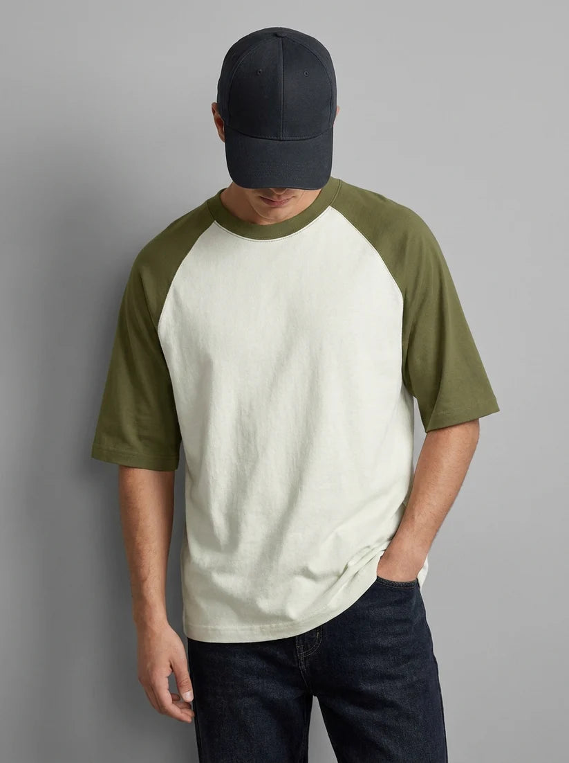 A man wearing a short-sleeve raglan t-shirt with an off-white body and olive green sleeves, paired with a dark navy baseball cap and dark jeans, standing against a solid gray background.