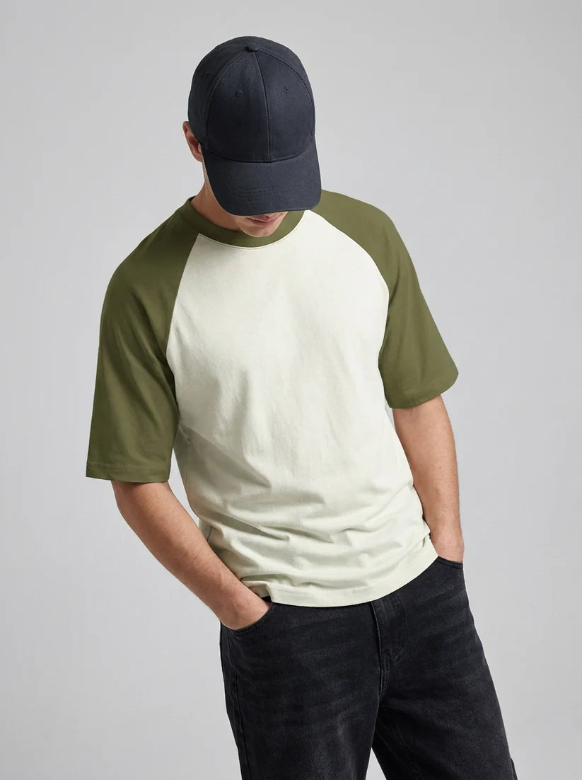 A man wearing a baseball tee with an off-white body and olive green raglan sleeves, paired with a dark navy cap and dark jeans, photographed against a neutral light gray background.