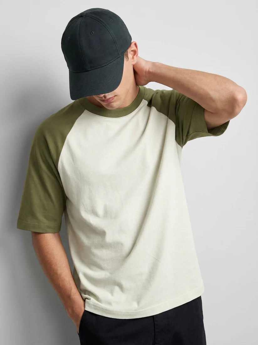 A man wearing a two-tone raglan t-shirt with an off-white body and olive green sleeves, paired with a dark green baseball cap and dark pants, posing against a light gray studio background.