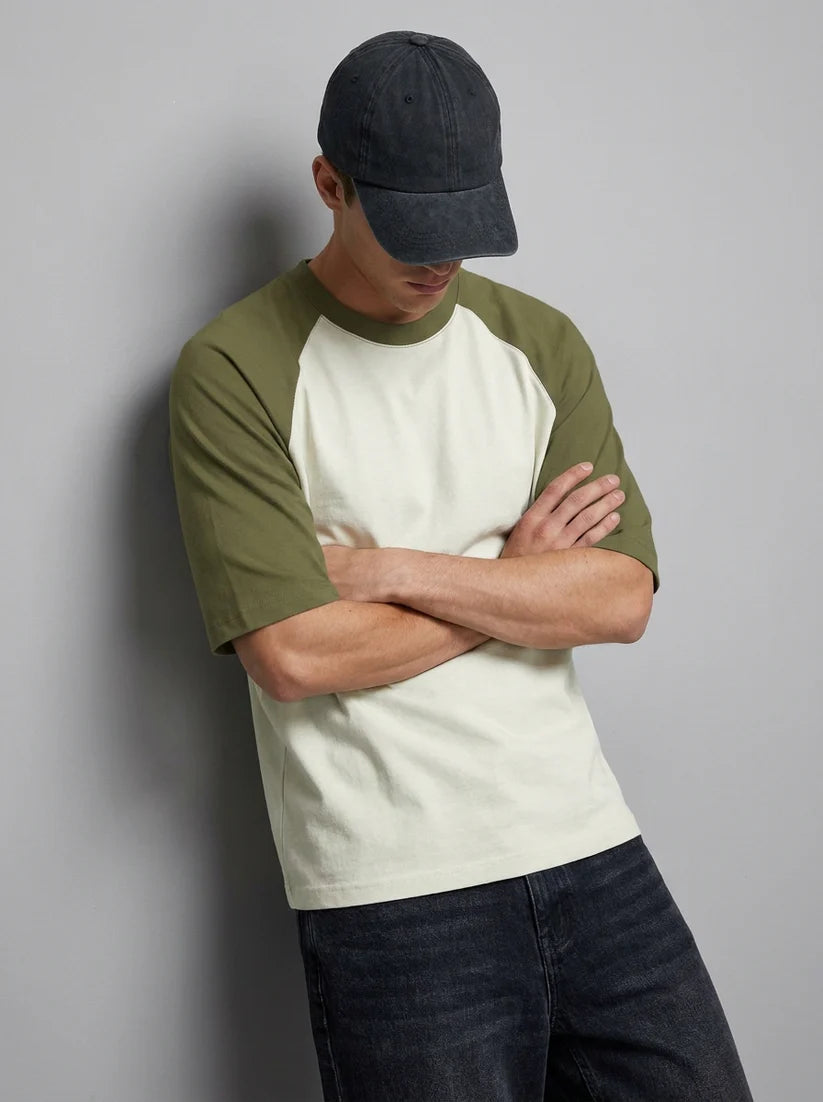 A man wearing a raglan-style t-shirt with an off-white body and olive green sleeves, paired with a dark gray baseball cap and dark jeans, leaning against a gray wall.