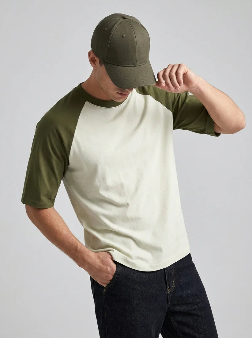 A man wearing a color-blocked raglan t-shirt with an off-white body and olive green sleeves, paired with a matching olive green baseball cap and dark jeans, against a solid light gray background.