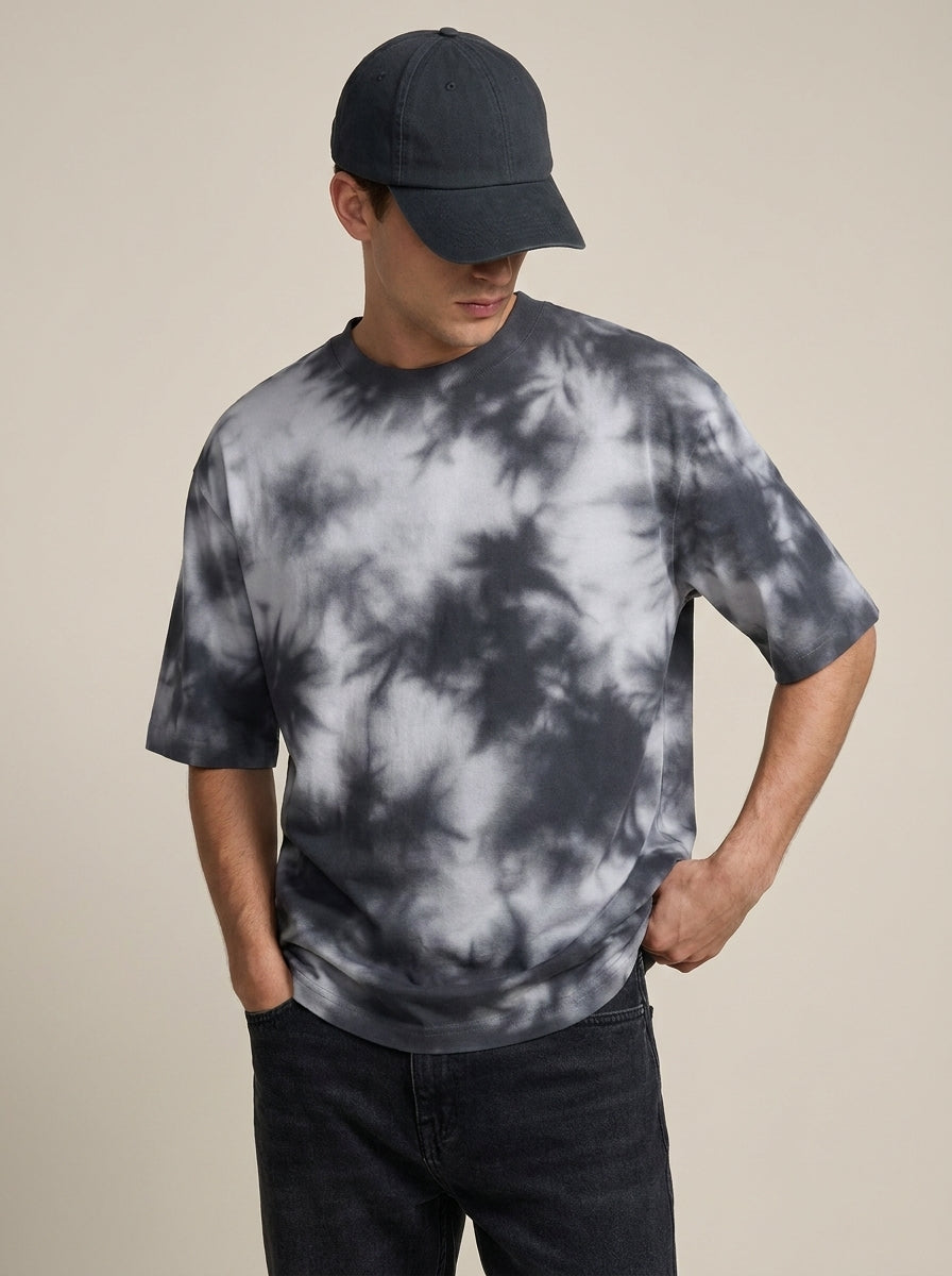 A man wearing a dark gray and white tie-dye t-shirt, a dark baseball cap, and dark jeans, standing against a solid beige background.