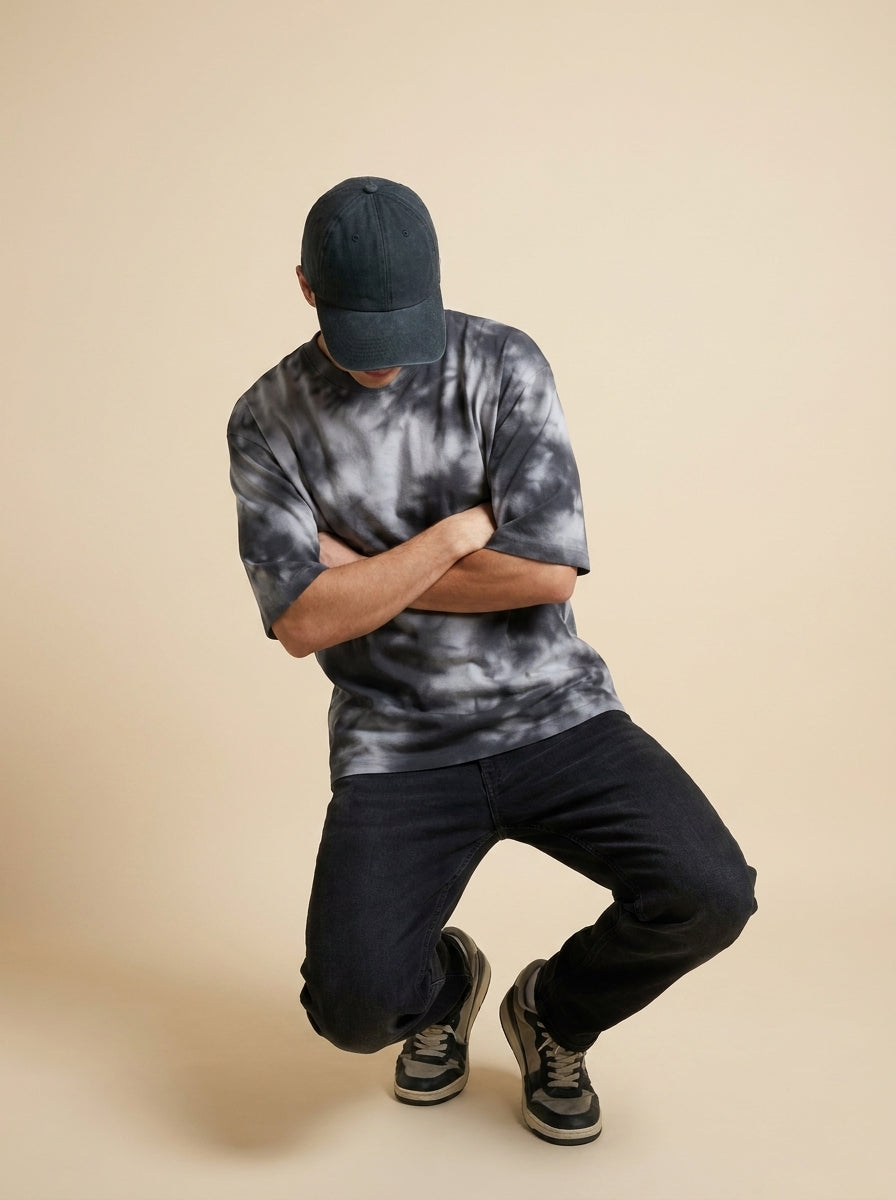 A man wearing a dark gray and black tie-dye t-shirt, dark jeans, and a black baseball cap, crouching in a dynamic pose against a solid beige studio background.
