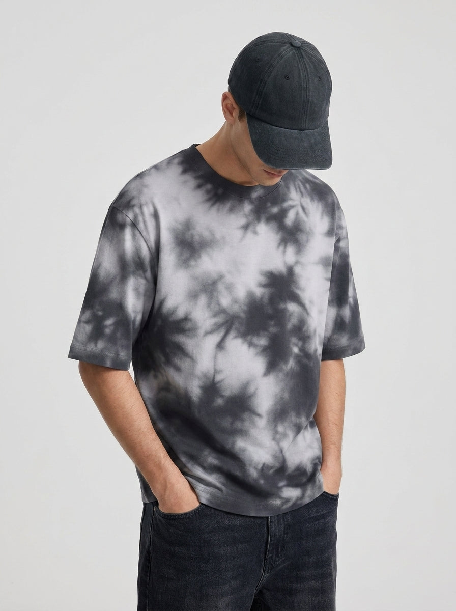 A man wearing a gray and white tie-dye t-shirt, a dark baseball cap, and dark jeans, standing against a light gray studio background.