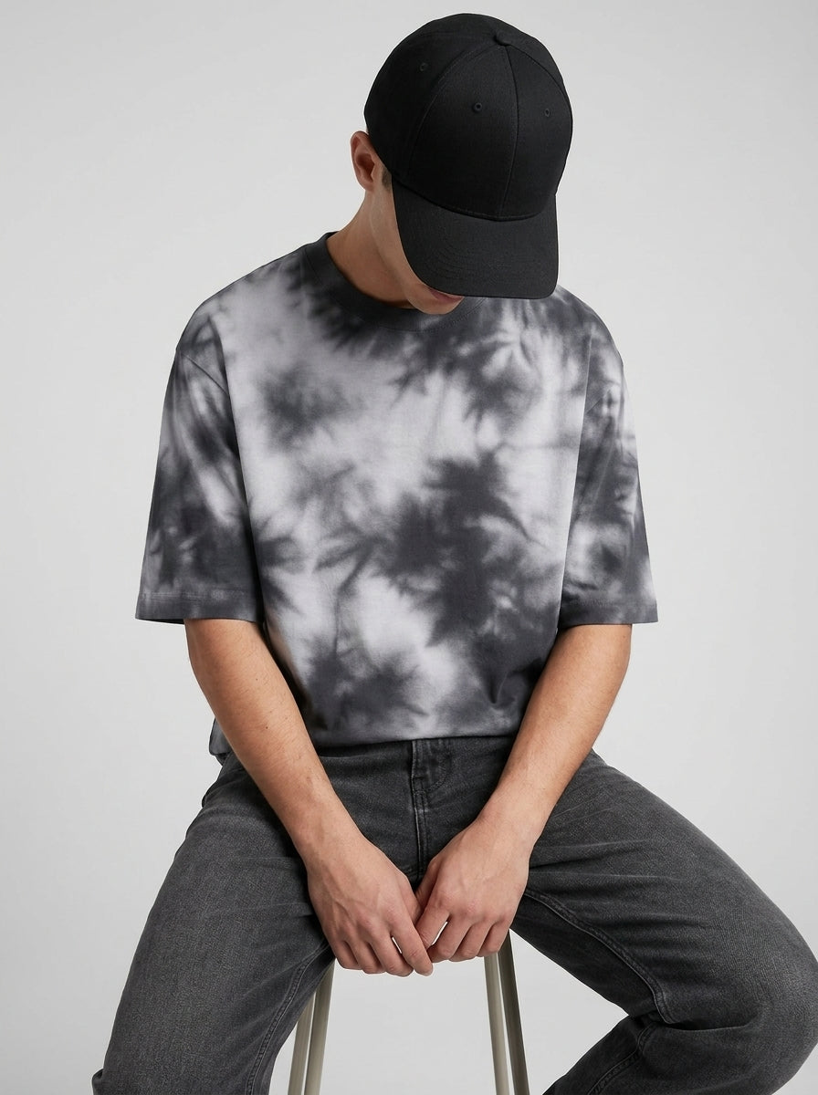 A man wearing a black baseball cap and a black and white tie-dye t-shirt, sitting on a stool against a light gray background.