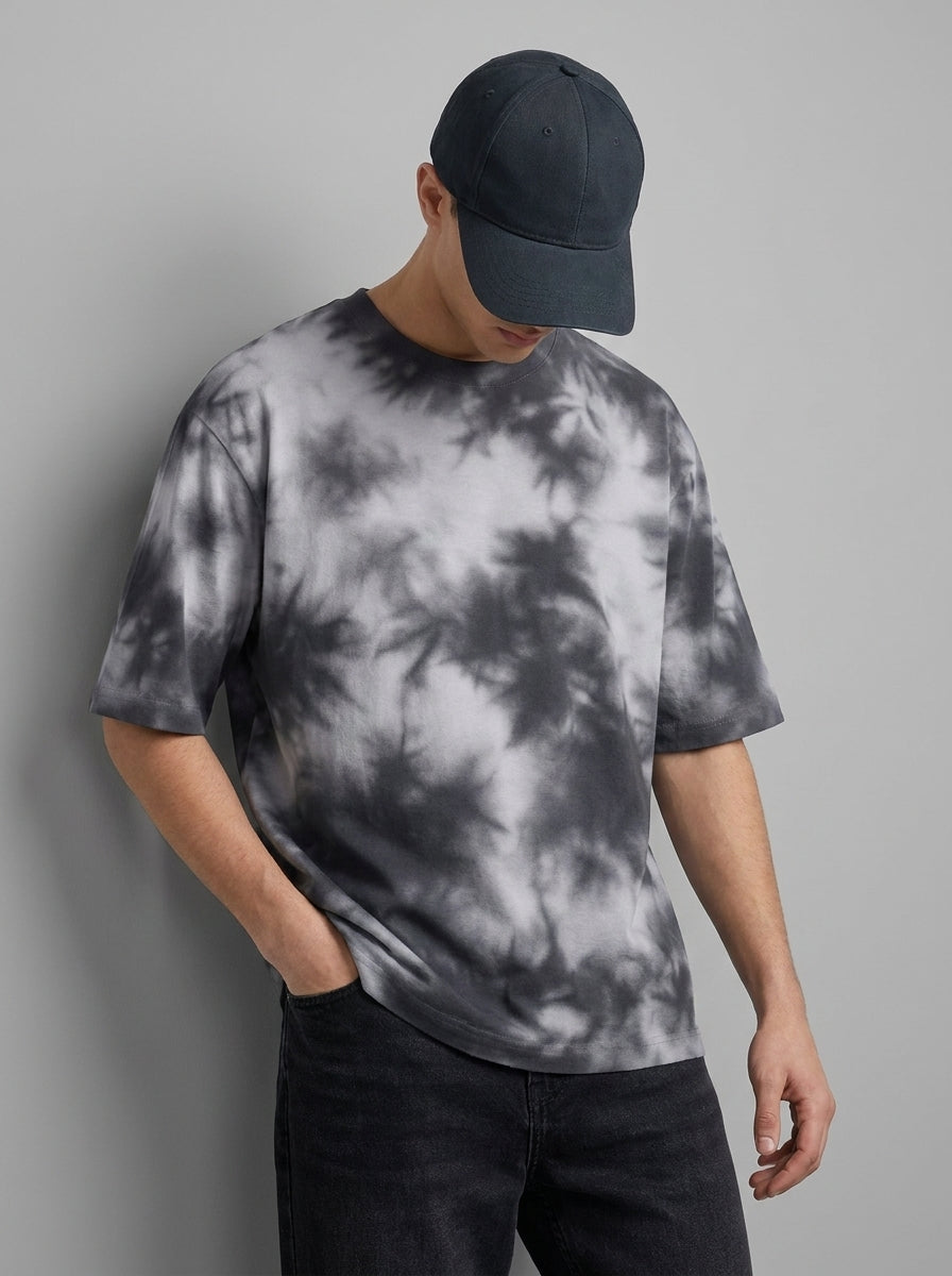 A man wearing a gray and white tie-dye t-shirt, a dark navy baseball cap, and dark jeans, standing against a solid gray studio background.
