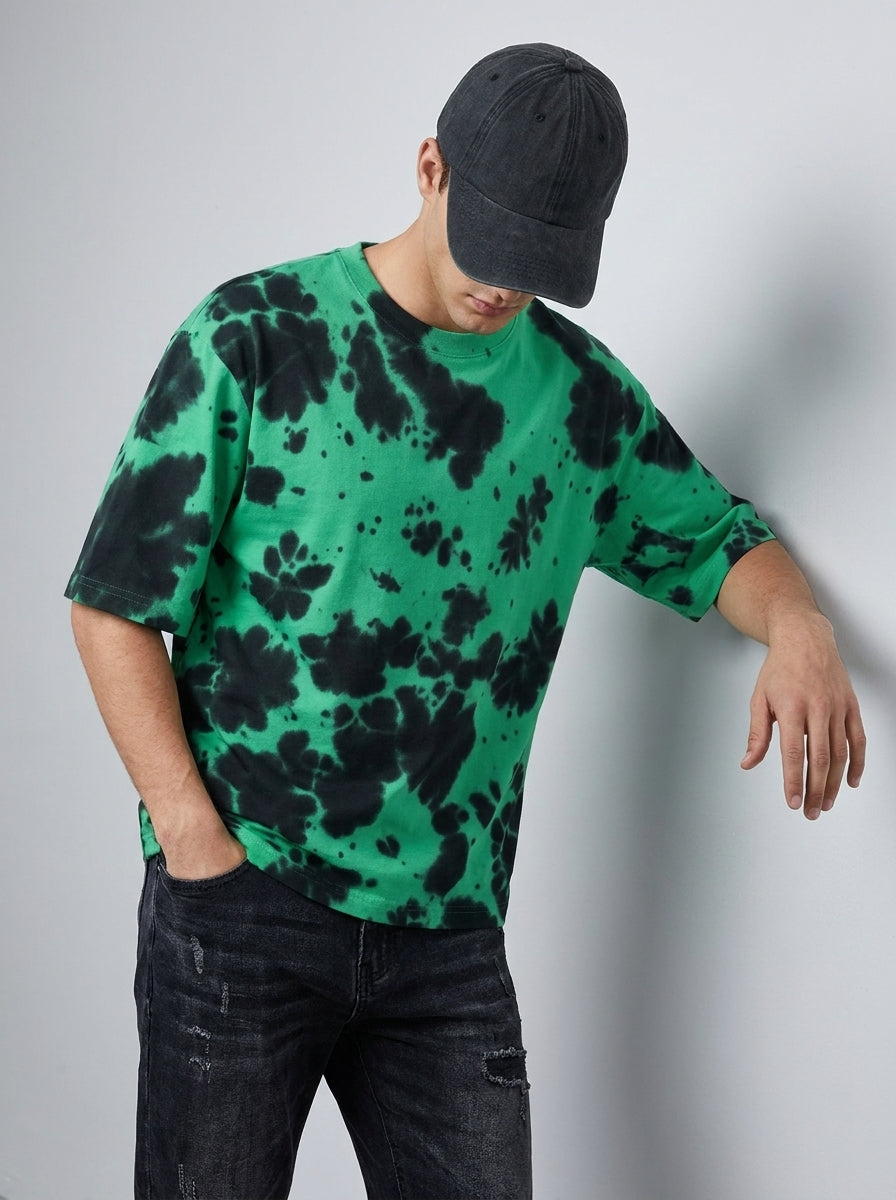 A man wearing a green and black tie-dye t-shirt, a black baseball cap, and dark distressed jeans, leaning against a light gray wall.