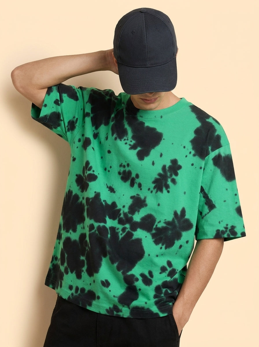 A man wearing a green short-sleeve t-shirt with a black tie-dye pattern, a black baseball cap, and black pants, posing against a solid beige background.