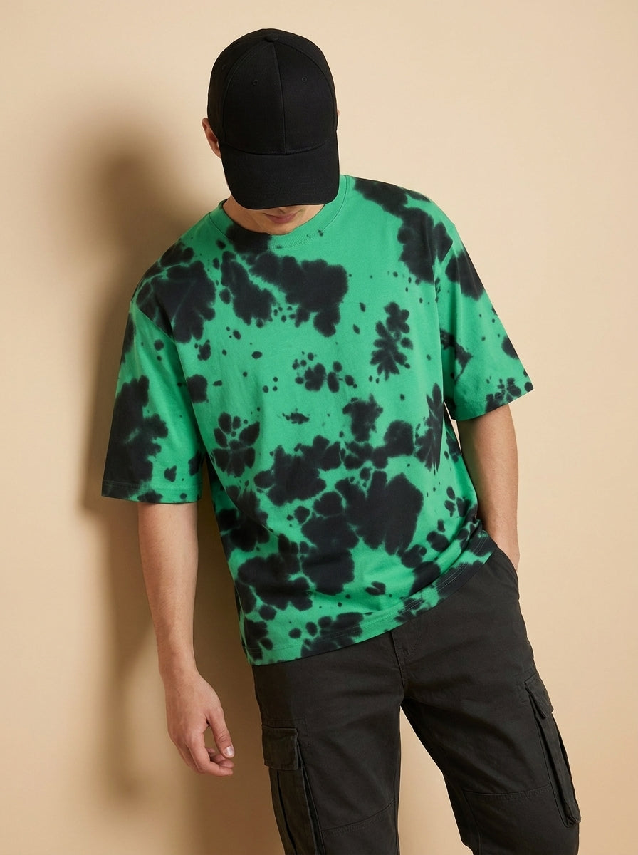 A man wearing a green and black tie-dye t-shirt, black cargo pants, and a black baseball cap, leaning against a beige wall.