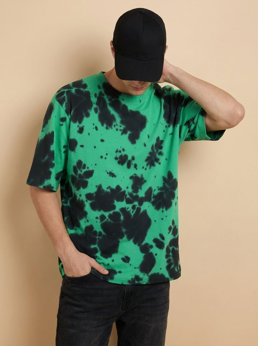 A man wearing a green short-sleeve t-shirt with a black tie-dye pattern, a black baseball cap, and black jeans, posing against a solid beige background.