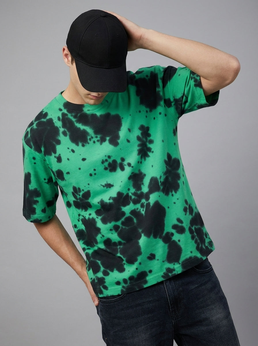 A man wearing a green short-sleeve t-shirt with a black tie-dye pattern, a black baseball cap, and dark jeans, posing against a solid gray studio background.
