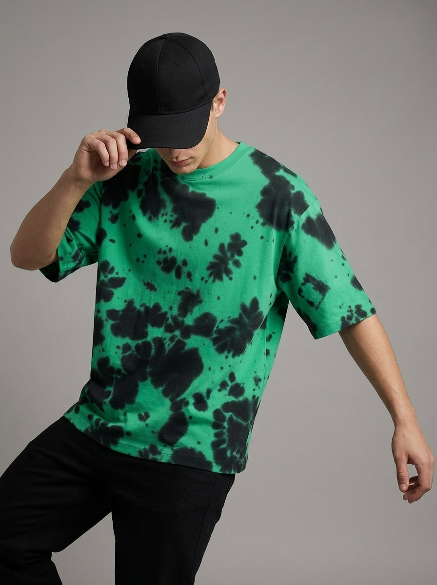 A man wearing a green and black tie-dye t-shirt, black pants, and a black baseball cap, posed against a solid gray studio background.