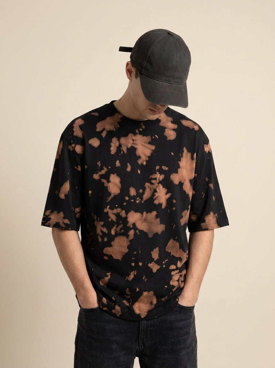 A man wearing a black short-sleeve t-shirt with a brown tie-dye pattern, a dark gray baseball cap, and dark jeans, standing against a solid beige background.