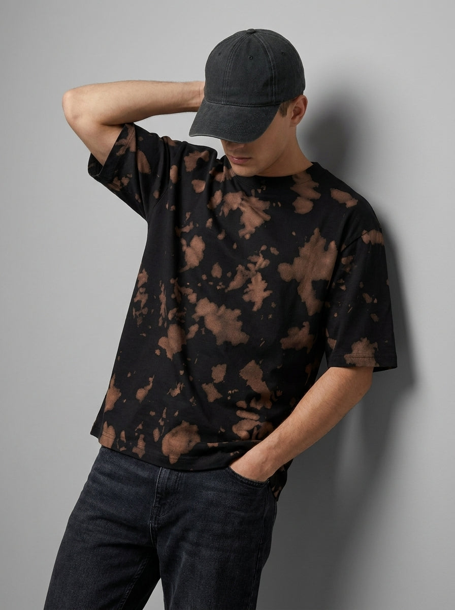 A man wearing a black short-sleeve t-shirt with a brown tie-dye pattern, a dark gray baseball cap, and dark jeans, leaning against a gray wall.