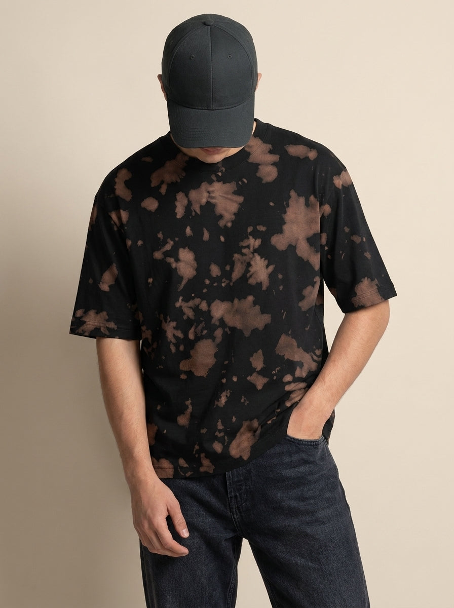 A man wearing a black short-sleeve t-shirt with a brown tie-dye pattern, a dark gray baseball cap, and dark blue jeans, standing against a solid beige background.