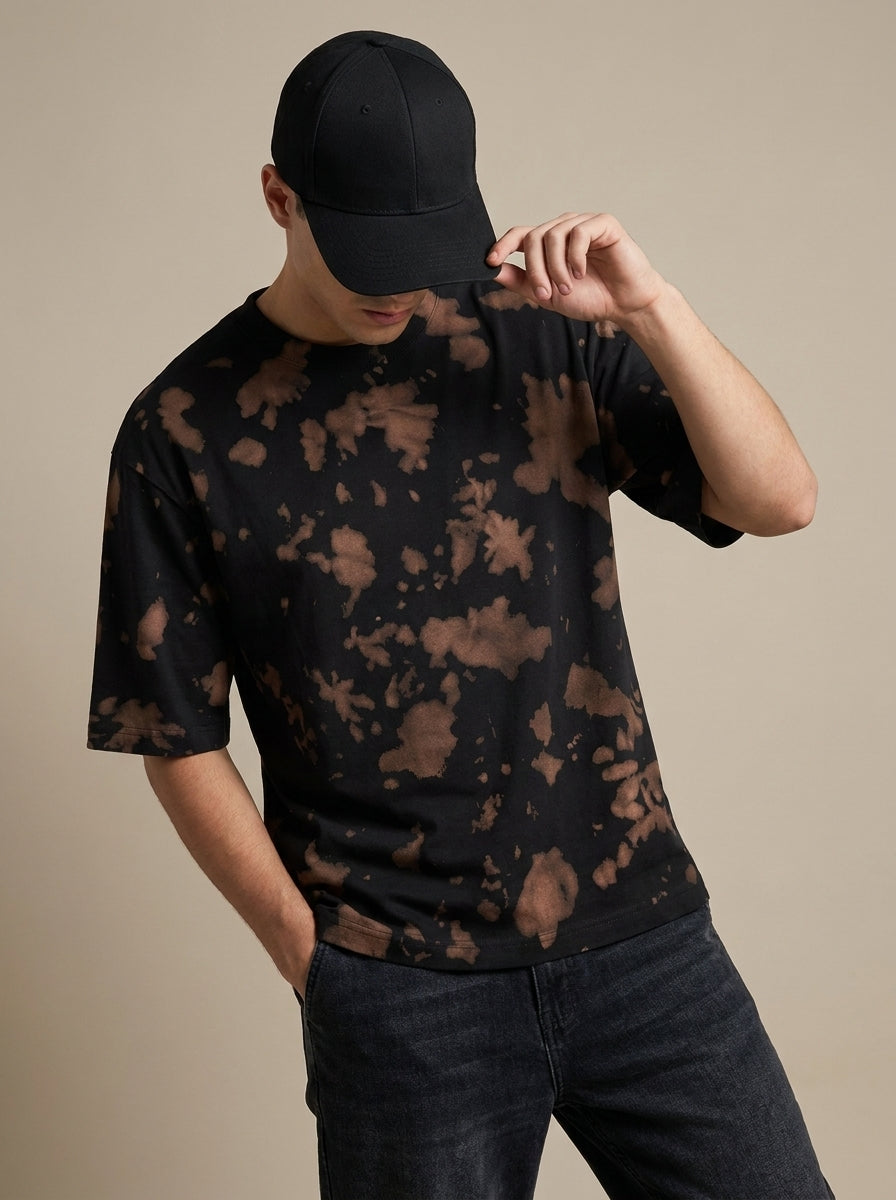 A man wearing a black short-sleeve t-shirt with a brown tie-dye pattern, a black baseball cap, and dark jeans, posing against a solid beige background.