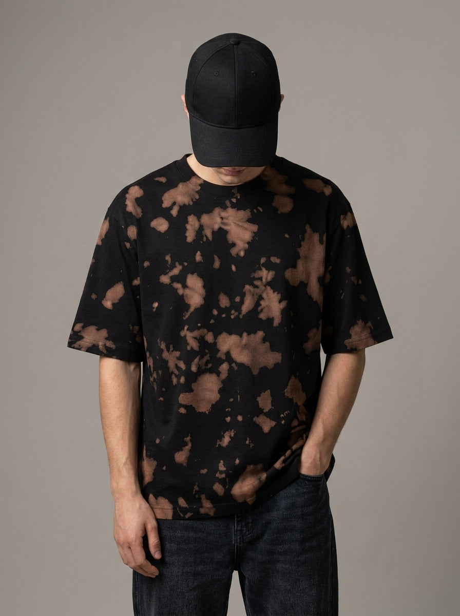 A man wearing a black short-sleeve t-shirt with a brown tie-dye pattern, a black baseball cap, and dark jeans, standing against a solid gray studio background.