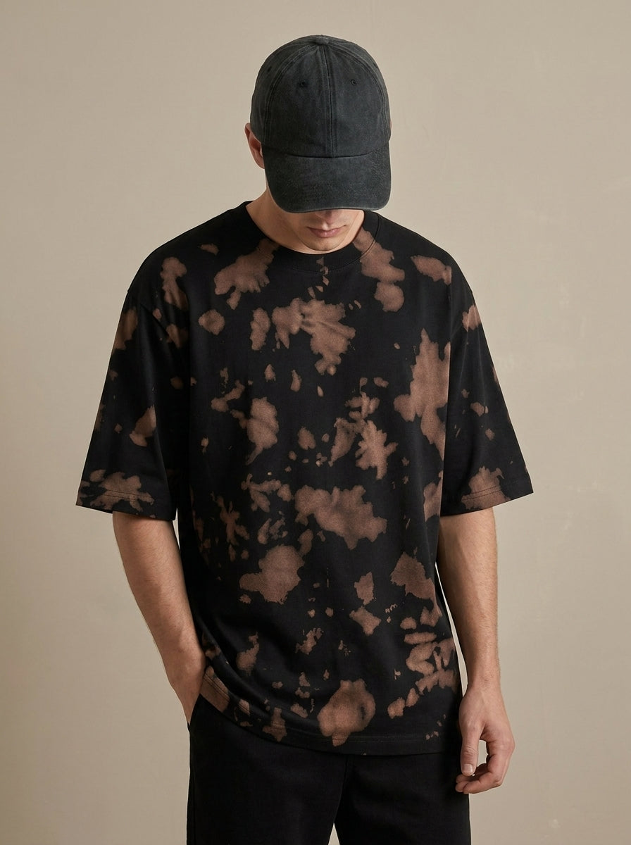 A man wearing a black short-sleeve t-shirt with a brown tie-dye pattern, a dark gray baseball cap, and black pants, standing against a plain beige background.