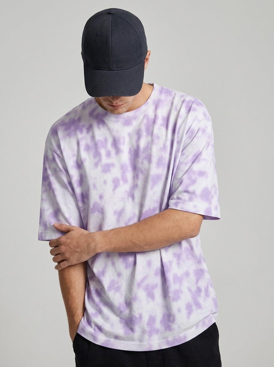 A man wearing a light purple and white tie-dye t-shirt, a black baseball cap, and black pants, standing against a solid light gray background.