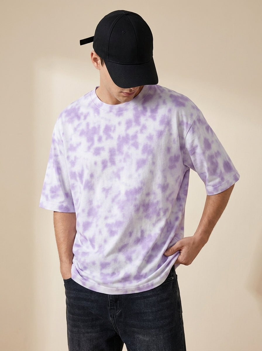 A man wearing a white short-sleeve t-shirt with a light purple tie-dye pattern, paired with a black baseball cap and dark jeans, standing against a solid beige background.