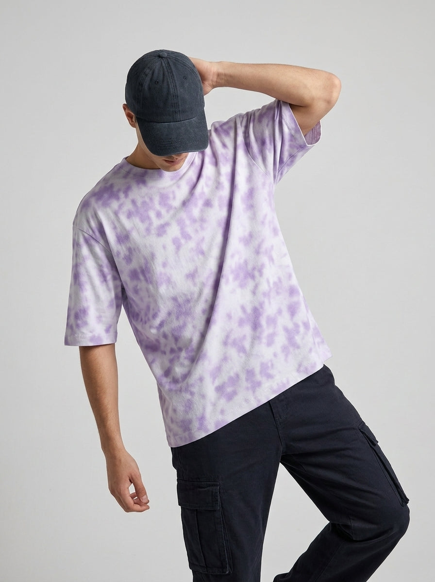 A man wearing a light purple and white tie-dye t-shirt, a dark navy baseball cap, and dark cargo pants, posing against a solid light gray background.
