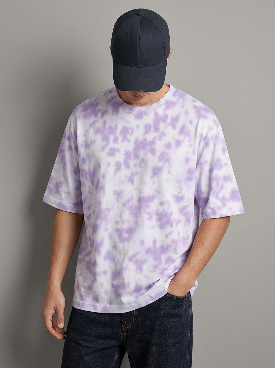 A man wearing a short-sleeve crewneck t-shirt with a light purple and white tie-dye pattern, paired with a dark navy baseball cap and dark blue jeans, standing against a solid gray background.