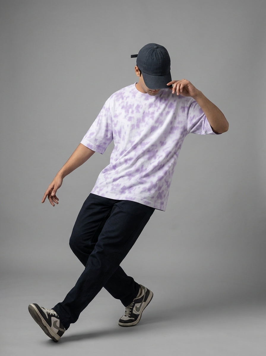A man wearing a light purple and white tie-dye t-shirt, dark navy pants, a dark navy baseball cap, and sneakers, captured in a dynamic pose against a solid gray studio background.