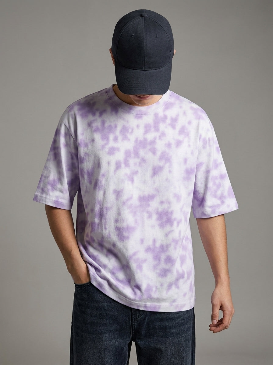 A man wearing a lavender and white tie-dye t-shirt, a dark navy baseball cap, and dark jeans, standing against a solid gray studio background.